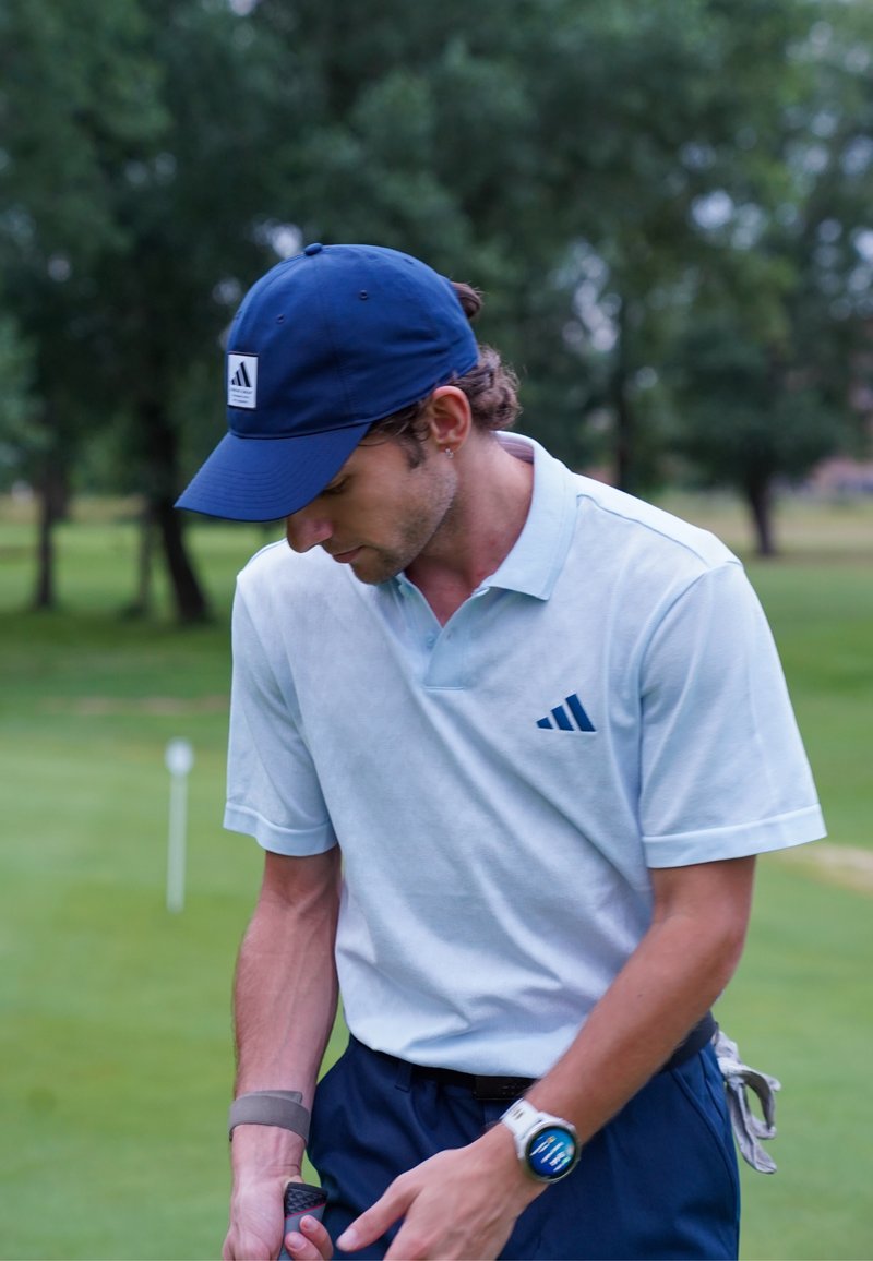 Mann med blå caps og hvit polo holder en golfkølle på en grønn golfbane med trær i bakgrunnen.