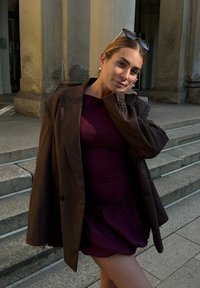 Brązowa oversize'owa marynarka na burgundowej sukience z rozkloszowanym dołem. W tle widać kamienne schody i kolumny. Okulary przeciwsłoneczne leżą na marynarce.
