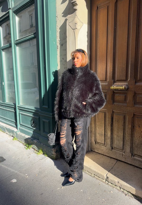 Γυναίκα με μαύρο γούνινο μπουφάν και σκισμένο μαύρο παντελόνι στέκεται σε ηλιόλουστο δρόμο δίπλα σε πράσινο παράθυρο και ξύλινη πόρτα, κρατώντας μια μικρή μαύρη τσάντα.