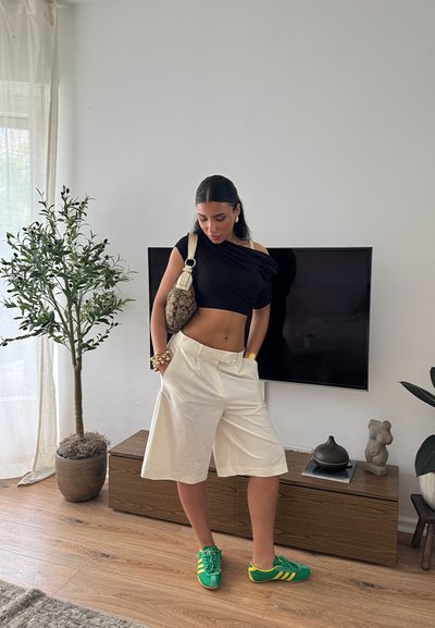 Top corto negro de hombros descubiertos, pantalones cortos blancos de pierna ancha, zapatillas verdes y amarillas, bolsa de patrón tostado, accessorios con pulseras.