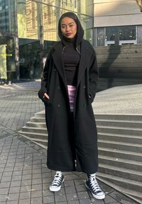 Schwarzer Wollmantel mit Kapuze, doppelreihigem Design, getragen über einem schwarzen Oberteil und einem Batik-Rock. Schwarz-weiße High-Top-Sneaker. Hintergrund aus quadratischem Pflaster.