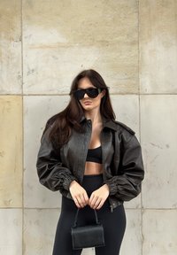 Mujer con chaqueta de cuero oscura, top corto negro, leggings negros, gafas de sol grandes y sosteniendo un pequeño bolso negro frente a una pared de piedra.