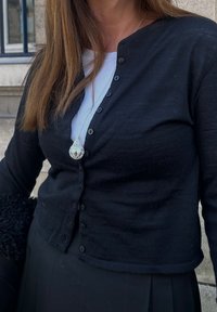Črn rebrast cardigan s skrajšano zasnovo in gumbi spredaj, oblečen preko bele srajce. Opazna srebrna ogrlica s kapljičastim obeskom.