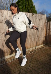 Femme portant une veste blanche et un legging noir sautant à la corde à l'extérieur près d'une clôture en bois par une journée ensoleillée.