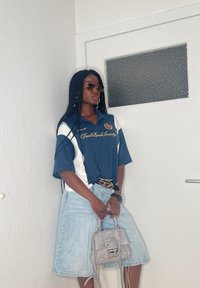 Blau-weißes, übergroßes Poloshirt mit dem Schriftzug "Fast Lane Society", kombiniert mit einem hellen Denim-Rock bis zum Knie und einer grauen Handtasche. Mit einer Sonnenbrille accessoriert.