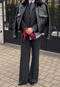 Personne portant un pantalon large à rayures fines, superposant des chemises foncées et claires, veste en cuir posée sur les épaules, tenant des gants en cuir rouge.