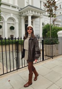 Jaqueta de couro castanha sobre um vestido de suéter bege, botas castanhas até o joelho, óculos de sol e um boné de tom claro, de pé perto de uma entrada com portão.