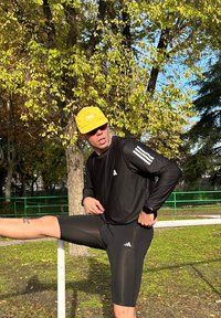 Camiseta deportiva negra de manga larga con tres rayas blancas, pantalones cortos ajustados negros y una gorra amarilla con logo. Estirándose contra una barra blanca.