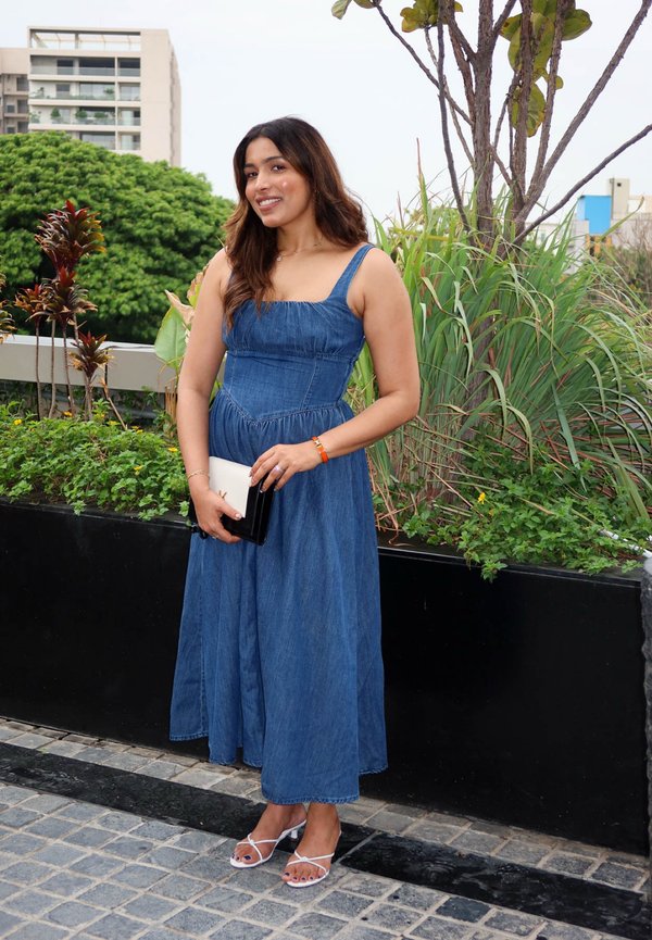 Frau in langem blauem ärmellosem Kleid und weißen Sandalen steht draußen lächelnd bei Pflanzen und hält eine kleine schwarz-weiße Clutch.