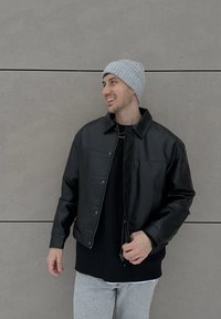 Homme portant un bonnet gris, une veste en cuir noire et un pull noir, debout devant un mur de béton gris, souriant et regardant vers la droite.