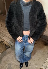 Personne portant une veste noire en tissu duveteux, un col roulé gris raccourci, un jean bleu ample, des chaussures noires pointues, et une bague en or à la main gauche.