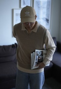 Beige knitted sweater with a polo collar, light blue jeans, and a tan Adidas cap. Holding magazines indoors with a dark sofa in the background.