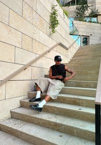 Μαύρο αμάνικο πλεκτό τοπ, μπεζ σορτς, λευκές κάλτσες και μαύρα παπούτσια. Καθισμένος σε πέτρινα σκαλιά με υφασμάτινο μπεζ τοίχο και φυ greenery.