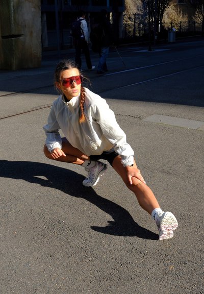 Figura atlética vestida con una chaqueta gris claro y shorts negros, estirándose. Lleva gafas de sol rosas y zapatillas blancas con suelas texturizadas.