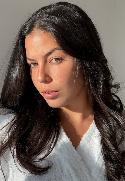 Joven con cabello largo y oscuro, ojos marrón claro y pecas, vestida con una prenda blanca texturizada, iluminada por luz natural sobre un fondo liso.