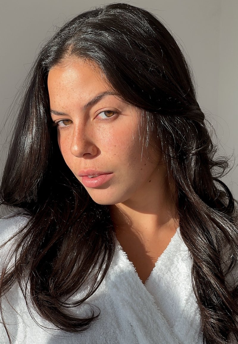 Joven con cabello largo y oscuro, ojos marrón claro y pecas, vestida con una prenda blanca texturizada, iluminada por luz natural sobre un fondo liso.
