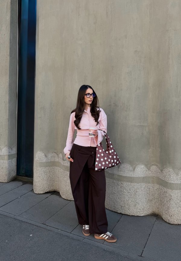 Femme aux longs cheveux bruns portant des lunettes, un haut rose, un pantalon large noir, tenant un sac à pois et un gobelet transparent, debout sur le trottoir en ville.
