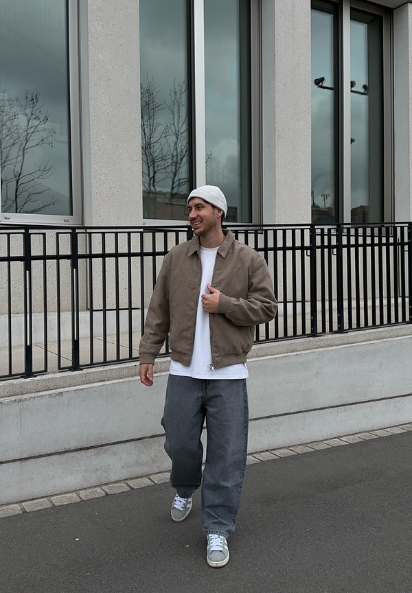 Moški v bež jakni, beli srajci, sivih hlačah in beli kapa beanie hodi po pločniku ob stavbi z velikimi okni in črnim ograjo.