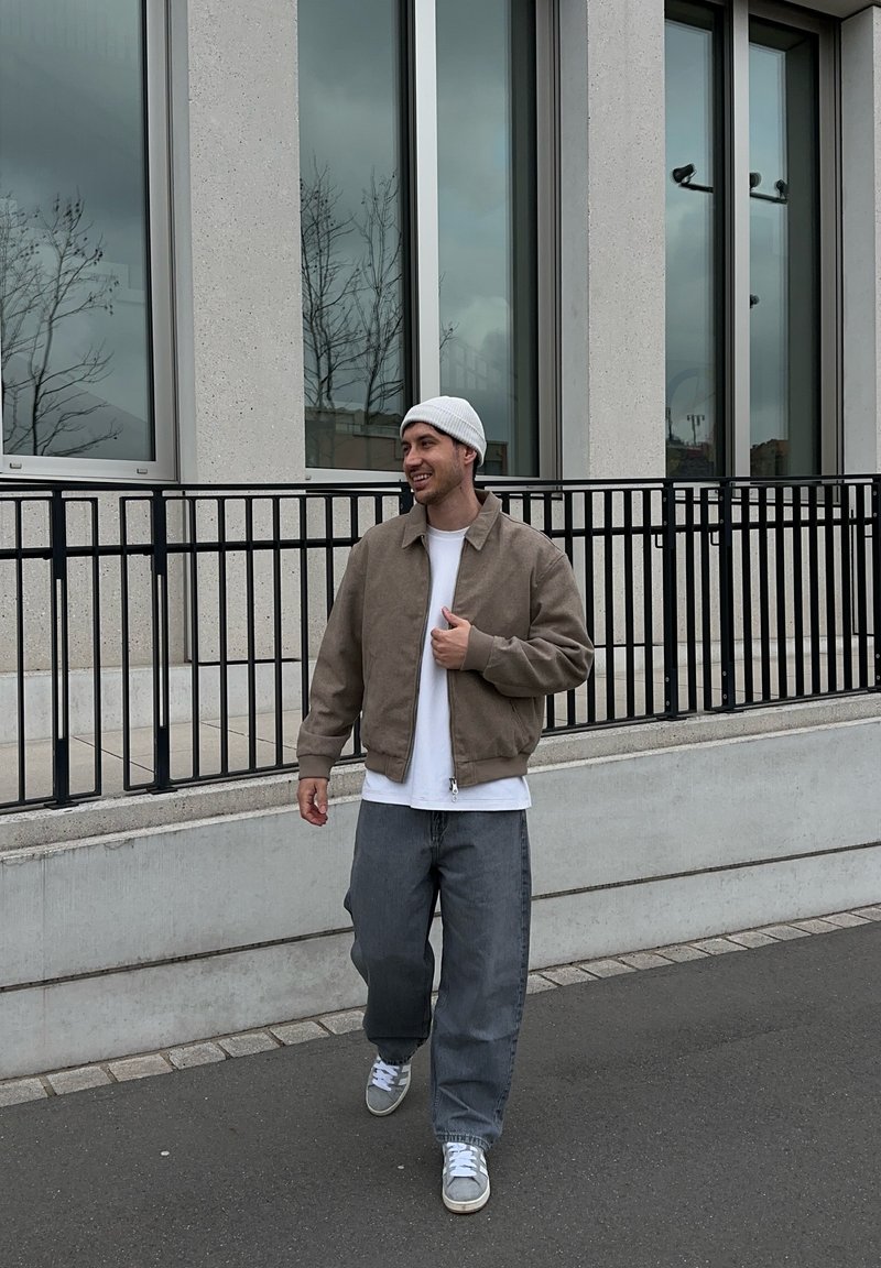 Vīrietis, tērpies bēša krāsas žaketē, baltā krekliņā, pelēkās biksēs un baltā cepurītē, iet pa ietvi blakus ēkai ar lieliem logiem un melnu margu.