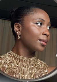 Złoty naszyjnik z warstwowymi splotami i perłowymi akcentami. Gładka skóra z dramatyczną kreską eyelinerem, odzwierciedlająca kobiecość w designie.