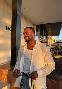 Valge linane särk valge põhitopiga, teksapüksid, käes päikeseprillid. Soe valgustus rõhutab kiviseina ja õueistumiskohta.