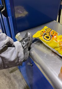 Scarpa da ginnastica nera con dettagli in rete poggia su una superficie metallica. Un sacchetto di snack Cheetos di un brillante colore giallo si trova nelle vicinanze.