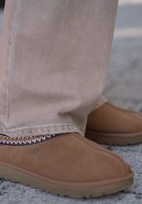 Καφέ σουέντ παντόφλες UGG φορεμένες με μπεζ παντελόνια σε επιφάνεια από χαλίκι, που δείχνουν λεπτομερή ραφές και καουτσούκ σό λες.