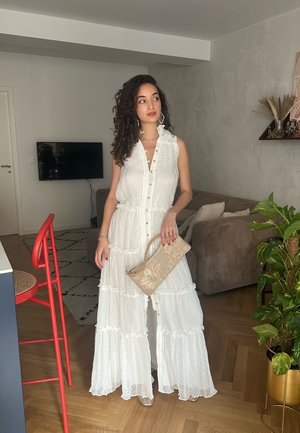 Vestido maxi blanco con volantes en capas, botonadura frontal y textura sutil. Combinado con un bolso de mano beige con patrones y una elegante silla de mimbre.