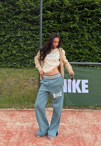 Mujer de pie en una pista de tenis roja, levantando su sudadera beige para mostrar su cintura, vestida con pantalones de deporte azules de Nike y sosteniendo unas gafas de sol.
