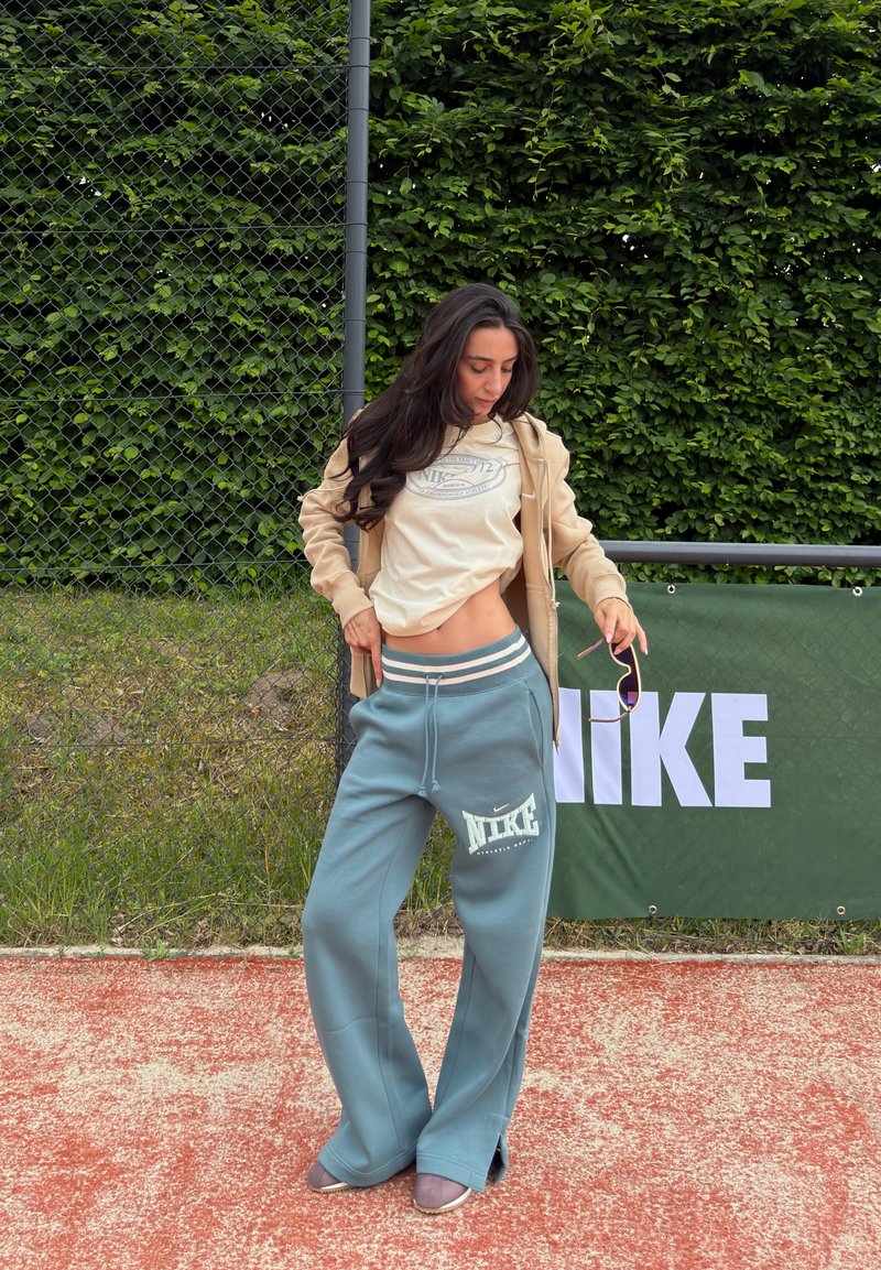 Femme debout sur un court de tennis rouge, soulevant son sweat-shirt beige pour montrer sa taille, portant un pantalon de survêtement bleu Nike et tenant des lunettes de soleil.