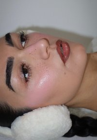 Profilansicht einer Person mit weißen Kunstfell-Ohrenschützern. Die Haut hat ein glattes, strahlendes Finish mit rosigen Wangen und glänzenden braunen Lippen. Braune Augen sind sichtbar.