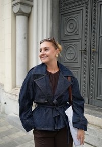 Džinsinė striukė su peplum dizainu, dideliais atvartais ir dirželiu juosmenyje. Dėvima ant tamsaus marškinėlio, derinama su balta rankine. Fone matoma tekstūruota duris.