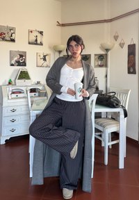 Szary długi kardigan na białym topie, do tego luźne ciemne spodnie w paski i beżowe botki. Trzyma kubek ze Starbucks, stoi w pomieszczeniu.