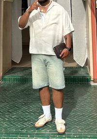 Hvitt kortermet skjorte, lyse denimshorts og beige sneakers med hvite sokker. Mørk crossbody-veske og tilbehør, inkludert ringer og halskjede.