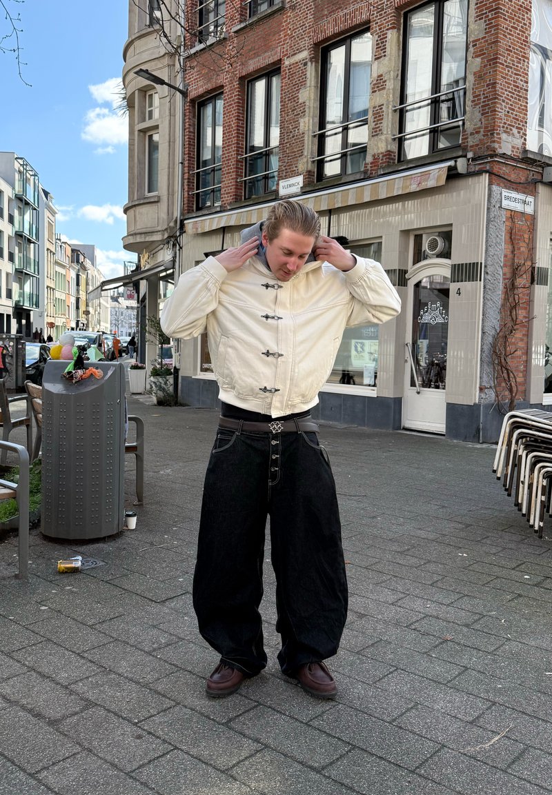 Un jeune homme ajuste la capuche de sa veste crème, debout sur le trottoir de la ville près d'un bâtiment en briques avec des panneaux de rue "Vleminck" et "Bredestraat".