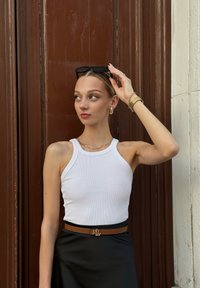 Débardeur blanc côtelé, jupe en satin noire, ceinture marron avec boucle en or, boucles d'oreilles dorées en créole, lunettes de soleil tenues à la main, fond neutre.