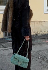 Sac à main matelassé vert menthe avec une bandoulière chaîne argentée et un fermoir logo proéminent. Positionné à côté d'une personne portant une tenue noire avec des accents en fourrure.