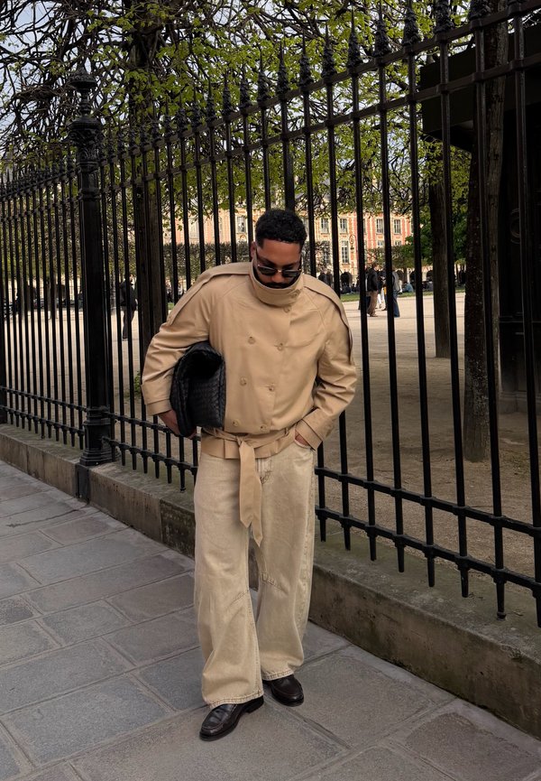Man draagt een beige trenchcoat en wijde broek, houdt een zwarte geweven clutch vast, loopt naast een hoog zwart metalen hek in een park.
