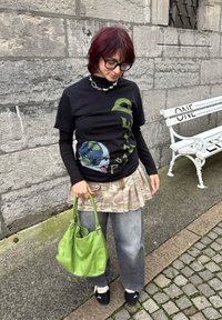 Crna grafička majica s dizajnom globusa, nošena ispod dugih rukava; siva suknja s maskirnim uzorkom; zelena torba; crne cipele s bijelim detaljima.