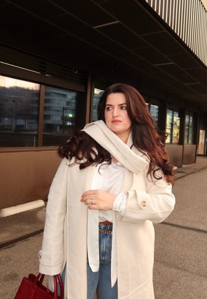 Mujer con cabello largo y oscuro, vestida con un abrigo crema y una bufanda, sosteniendo un bolso rojo, de pie frente a un edificio con ventanas reflectantes.
