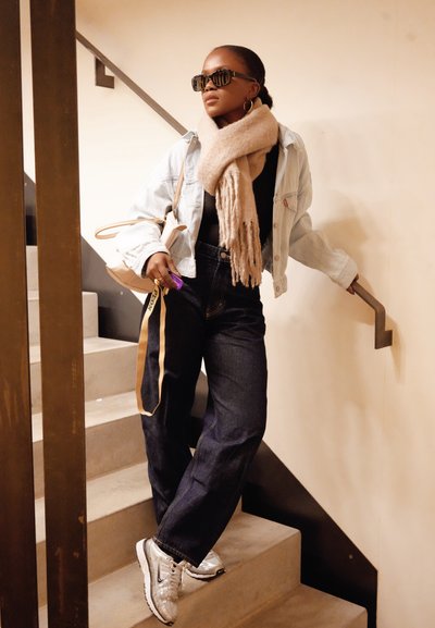 Chaqueta de mezclilla azul claro, camiseta negra, jeans oscuros de cintura alta, zapatillas plateadas, bufanda beige, gafas de sol negras, sosteniendo un bolso pequeño, fondo de escaleras.