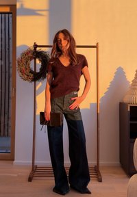 Femme portant un pull à manches courtes marron foncé et un jean large foncé, tenant une pochette sombre, avec une couronne décorative et un décor en bois en arrière-plan.