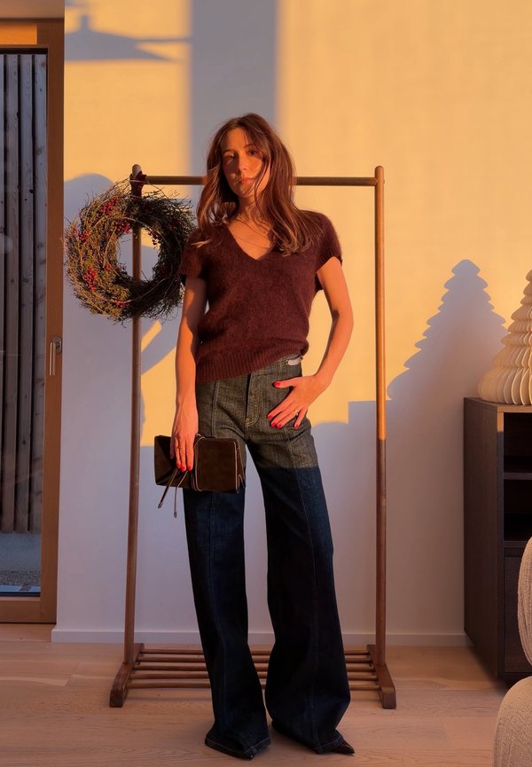 Frau in einem dunkelbraunen Kurzarm-Pullover und dunklen, weit geschnittenen Jeans steht, hält eine dunkle Clutch, im Hintergrund ist ein dekorativer Kranz und Holzdekoration zu sehen.