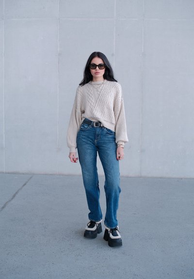 Mujer con cabello largo y oscuro, usando gafas de sol, suéter beige de punto trenzado, jeans azules y zapatos plataforma negros, de pie sobre suelo de concreto gris.
