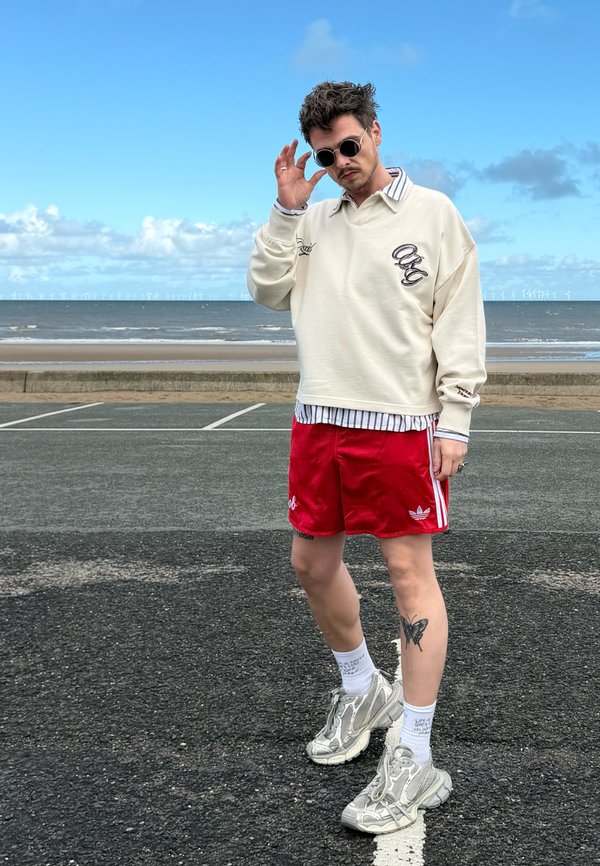 Person med solbriller, cremefarvet sweater, røde Adidas-shorts og grå sneakers står på asfalt nær stranden og justerer solbrillerne med den ene hånd.