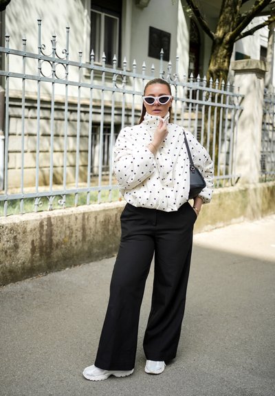 Mujer que lleva una chaqueta blanca con lunares, pantalones negros de pierna ancha, zapatillas blancas y gafas de sol de ojo de gato, está de pie en la calle cerca de una valla metálica.