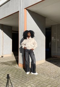 Jachetă bomber bej deschis, blugi evazați negri din denim cu logo pe lateral, adidași albi, stând îndreptat spre un perete texturat gri în lumina soarelui.