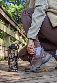Schwarze Wasserflasche mit weißen Illustrationen und Schraubverschluss, neben braunen Wanderschuhen mit türkisfarbenen Akzenten auf einem Holzsteg.
