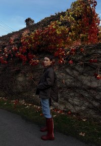 Prešívaná hnedá bunda, svetlomodré džínsy a vínové gumáky s bielymi akcentmi. V pozadí je kamenná stena a jesenné lístie.