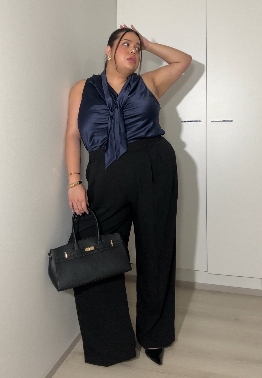 Femme portant un haut sans manches en satin bleu marine et un pantalon large noir, tenant un sac à main noir, posant avec une main sur la tête contre un mur blanc.